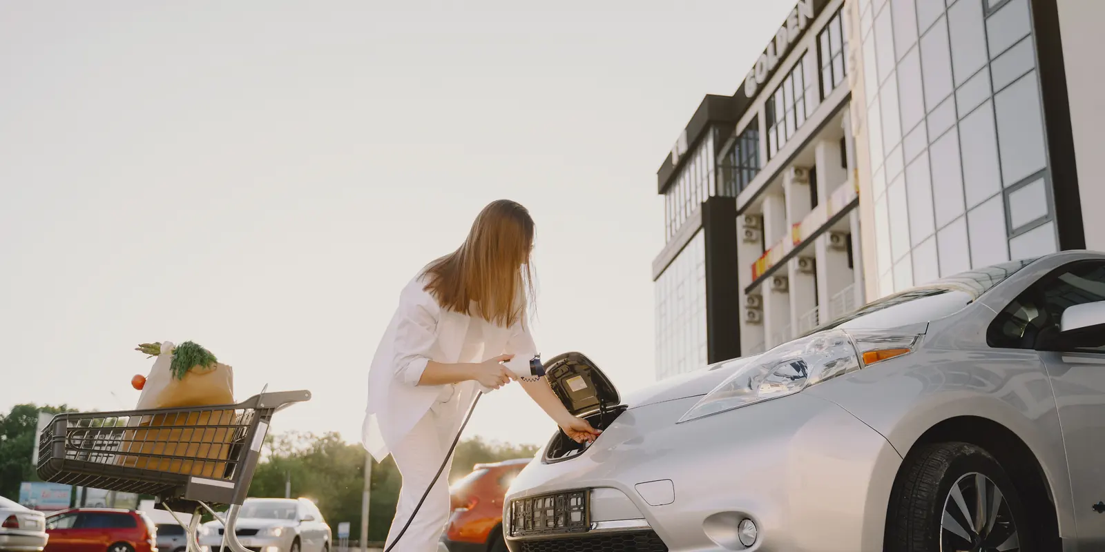 Nő elektromos autót tölt a bevásárlóközpont parkolójában, mellette teli bevásárlókocsi