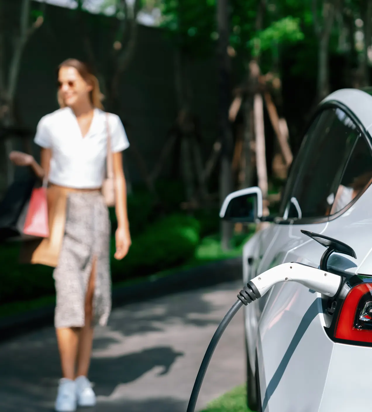Bevásárlótáskás nő sétál a töltőre csatlakoztatott fehér elektromos autó felé