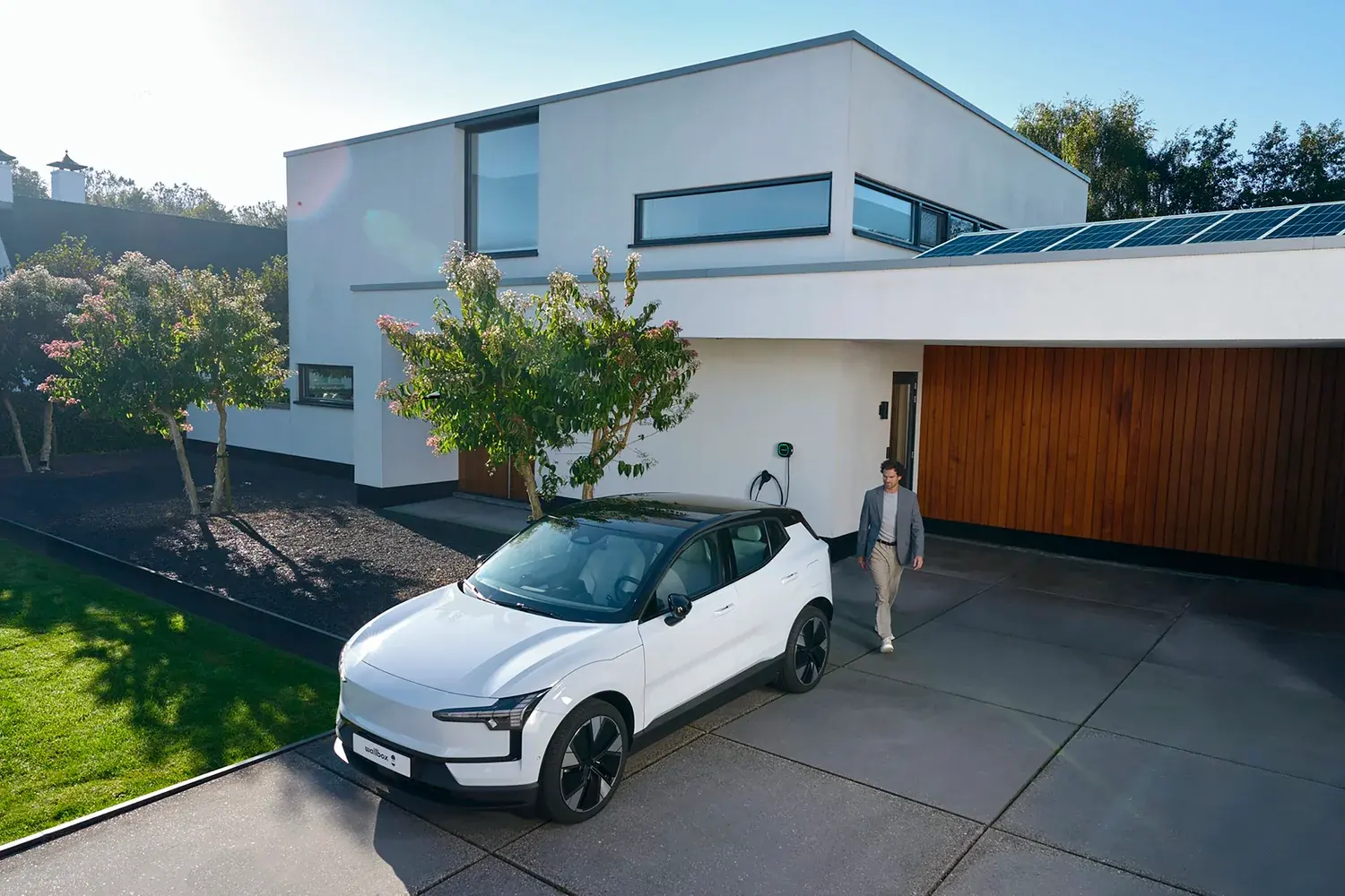 Férfi sétál a ház előtt parkoló elektromos autó mellett, a falon otthoni töltővel